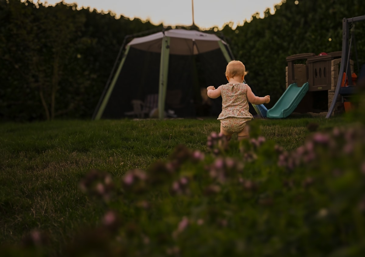 A baby in a floral romper walking across a grassy backyard at golden hour related to The Regalo My Cot Portable Toddler Bed: Comfort and Convenience on the Go