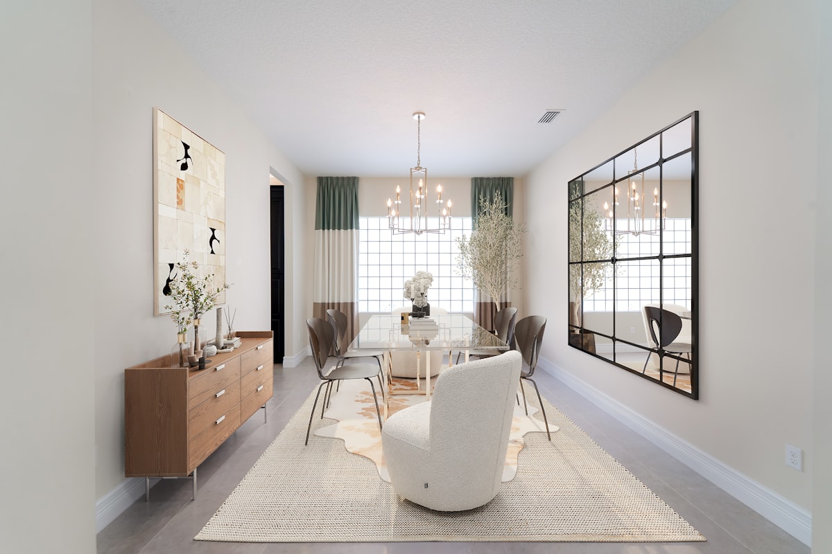 A bright and modern dining room featuring a glass-top table, neutral-toned chairs, and a cozy boucle accent chair. Natural light floods through a large glass block window, highlighting elegant greenery, a contemporary chandelier, and a large industrial-style mirror that adds depth and dimension. The soft color palette, sleek sideboard, and layered textures create a serene and stylish ambiance perfect for hosting or relaxing. related to Elevate Your Dining Experience: The Ultimate Guide to Sideboards and Buffets