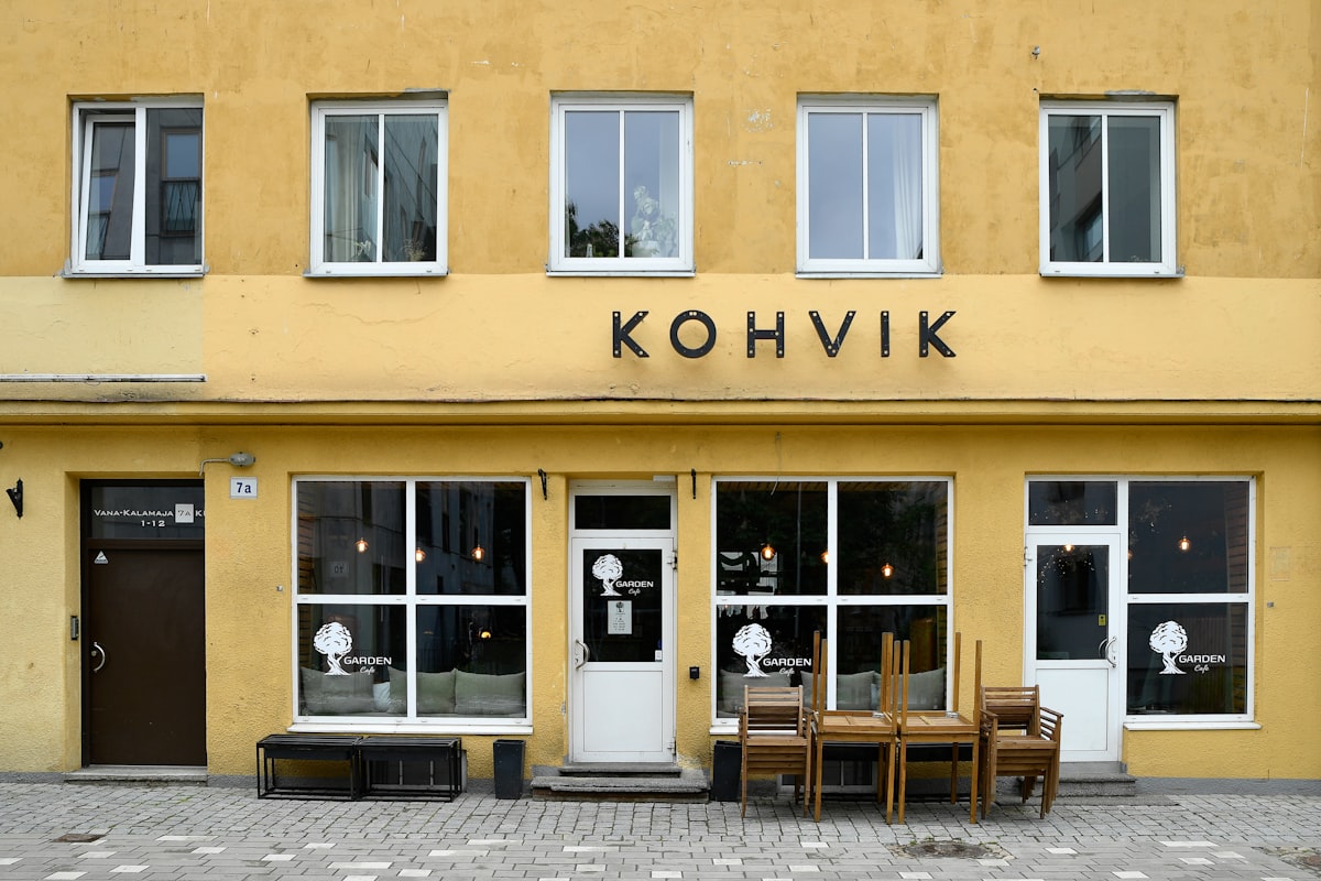 A charming and symmetric storefront of a traditional Estonian coffee shop with a vibrant yellow facade, featuring large glass windows, wooden outdoor furniture stacked up, and minimalist black signage reading "KOHVIK".📸 Photo by me IG: @margoevardson related to Welcome Home: Enhancing Your Entryway with Stylish Furniture
