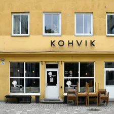 A charming and symmetric storefront of a traditional Estonian coffee shop with a vibrant yellow facade, featuring large glass windows, wooden outdoor furniture stacked up, and minimalist black signage reading "KOHVIK".📸 Photo by me IG: @margoevardson related to Welcome Home: Enhancing Your Entryway with Stylish Furniture