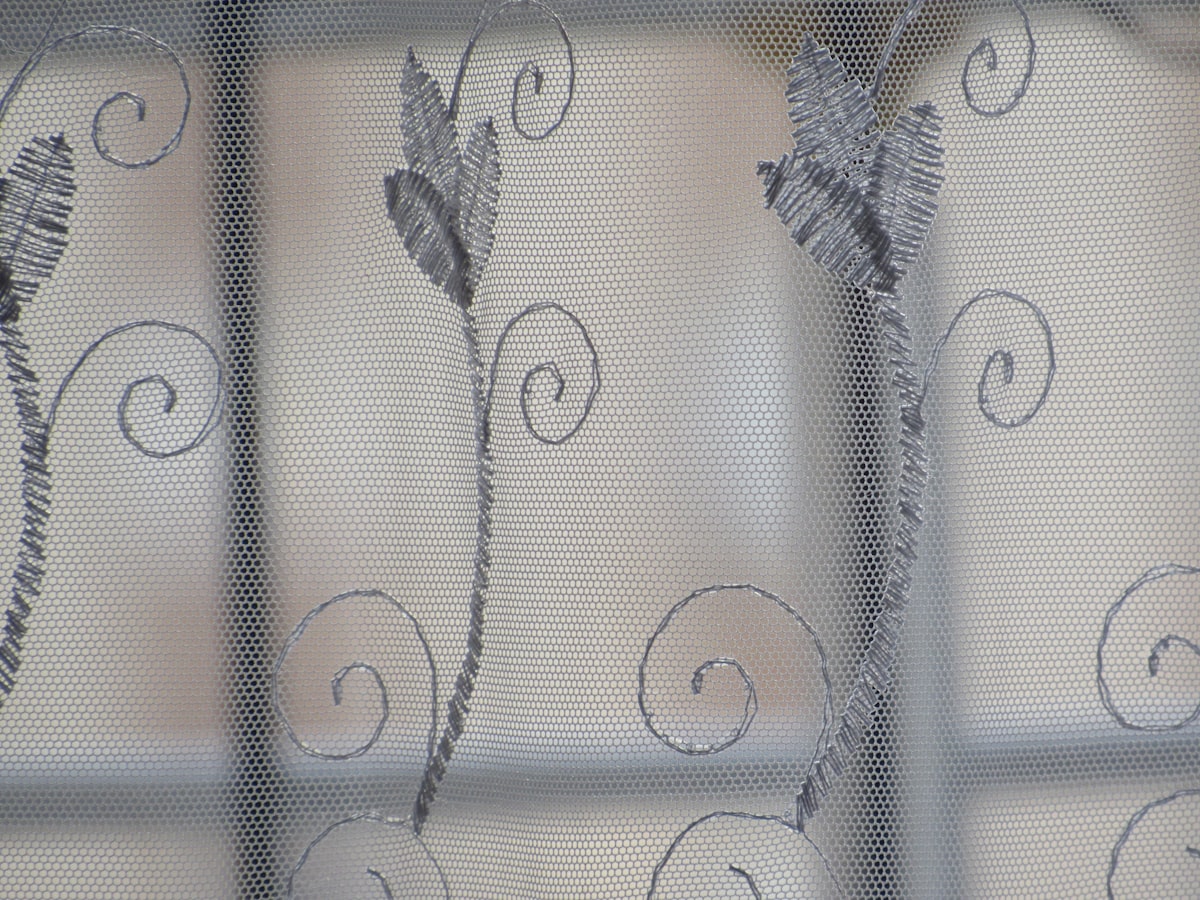 A close-up photo of a sheer curtain with a delicate floral pattern. The fabric is white and translucent, with light filtering through the weave. related to Window Treatments: Sheer Window Scarf Valances