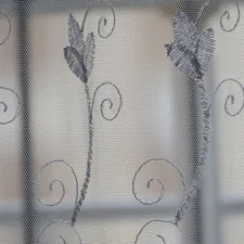 A close-up photo of a sheer curtain with a delicate floral pattern. The fabric is white and translucent, with light filtering through the weave. related to Window Treatments: Sheer Window Scarf Valances