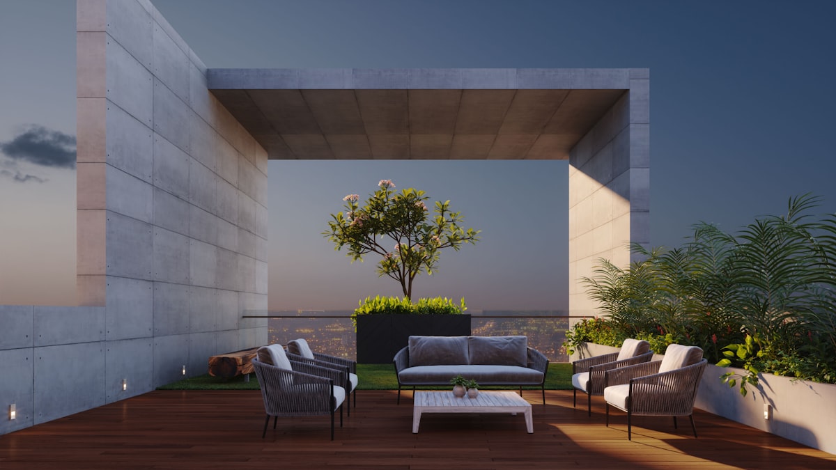 A minimalist architectural 3D rendering of a luxury rooftop terrace at dusk. The design is centered around a massive, open-frame brutalist concrete structure that perfectly frames a small, blossoming tree in a central planter. The seating area consists of a modern grey sofa and four matching woven armchairs arranged on a rich, dark wood deck, centered around a low-profile white coffee table. To the right, large built-in planters overflow with tropical ferns and greenery, while a small patch of artificial turf adds a touch of softness to the industrial aesthetic. In the background, the soft gl related to Enhance Your Outdoor Space with the Best Clay Chimineas at Lowes