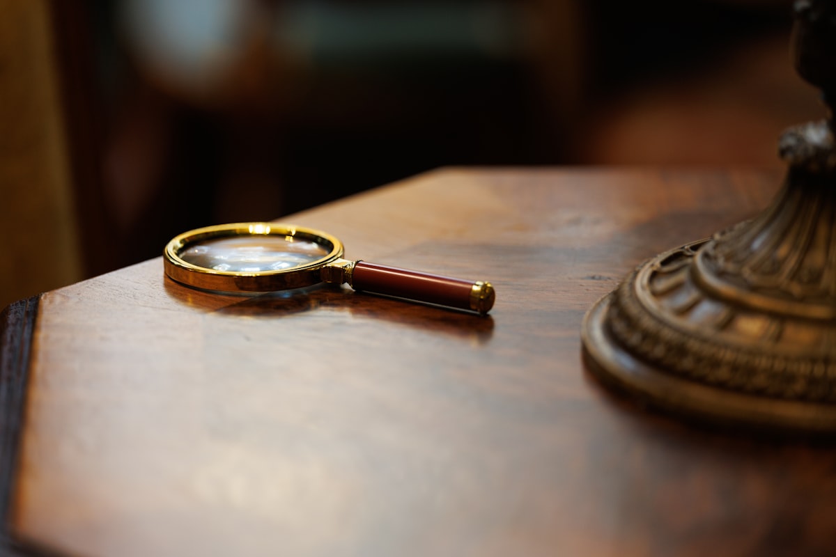 A small brass-rimmed magnifying glass with red handle rests on a worn wooden surface, with the gilded base of an antique lamp behind. Cozy detective-themed still life. related to Discovering Charm: A Closer Look at Waverly Garden Images Valance