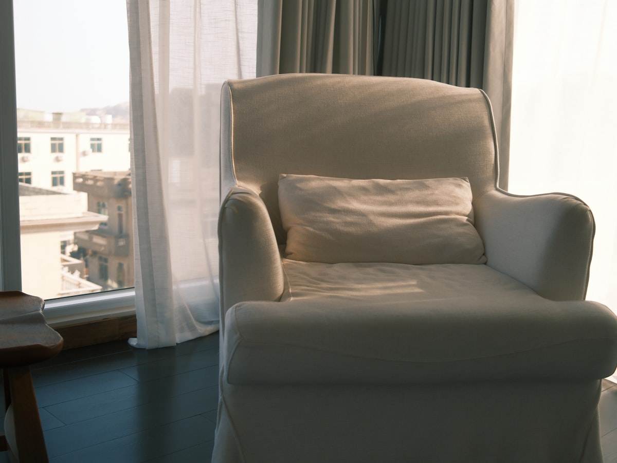 A white armchair with a pillow by the window. related to The Ultimate Space-Saving Solution: Chairs That Convert to Beds