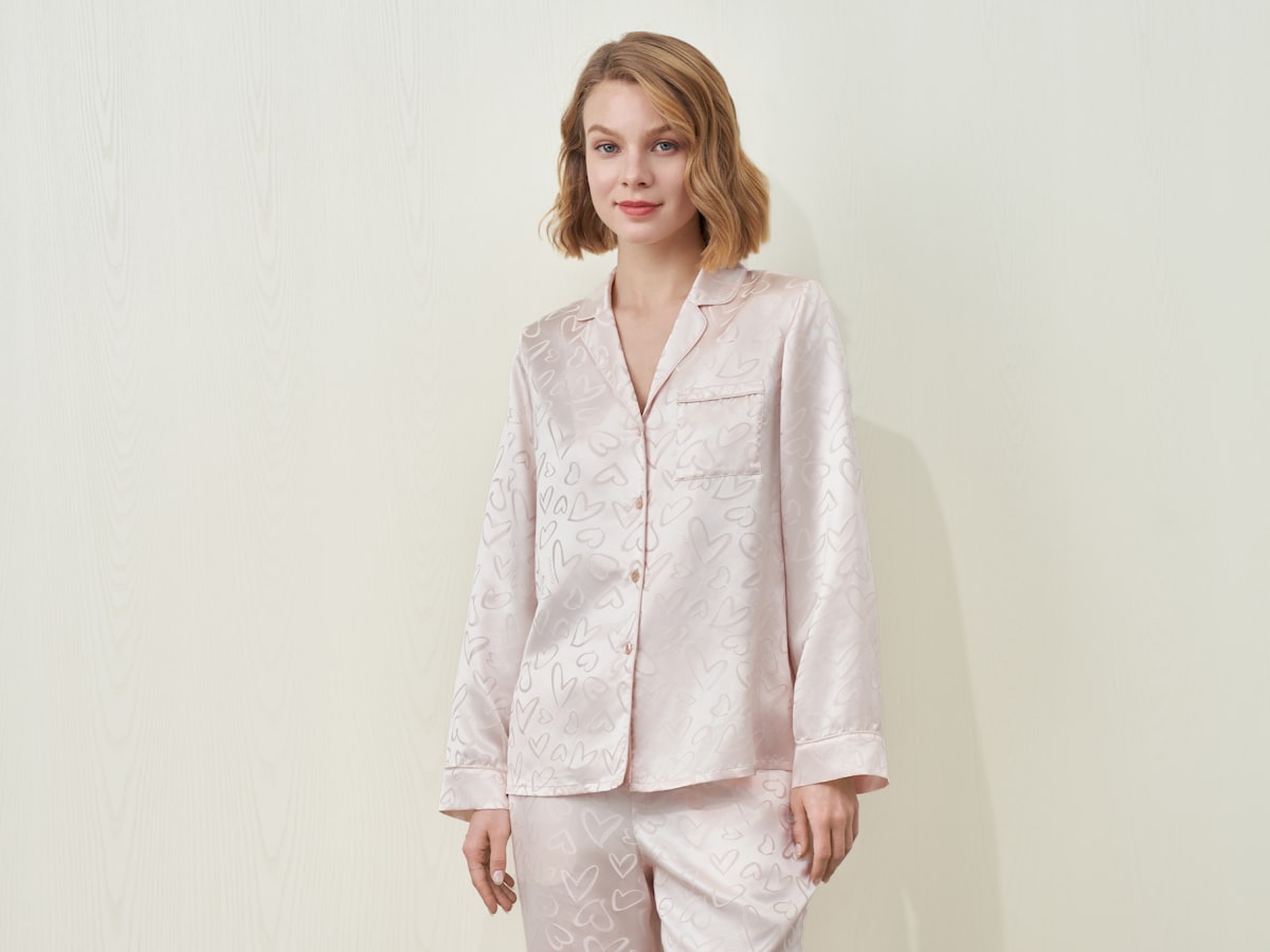 A woman in a pink MANITO silk pajama set with a subtle heart pattern against a studio wall, presenting soft sleepwear. related to Charming Bed Jackets for Women