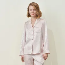 A woman in a pink MANITO silk pajama set with a subtle heart pattern against a studio wall, presenting soft sleepwear. related to Charming Bed Jackets for Women