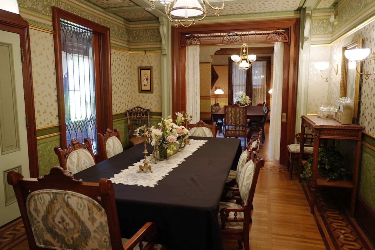 An elegant Victorian dining room at the Winchester Mystery House features carved wooden chairs, floral arrangements, and rich wallpaper with gold accents. The space flows into a second dining area, both lit by antique chandeliers and framed with lace curtains and intricate woodwork. This refined yet cozy setting contrasts the mansion's more mysterious features, showcasing its opulent domestic side. related to Dining Room Valance