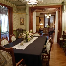 An elegant Victorian dining room at the Winchester Mystery House features carved wooden chairs, floral arrangements, and rich wallpaper with gold accents. The space flows into a second dining area, both lit by antique chandeliers and framed with lace curtains and intricate woodwork. This refined yet cozy setting contrasts the mansion's more mysterious features, showcasing its opulent domestic side. related to Dining Room Valance