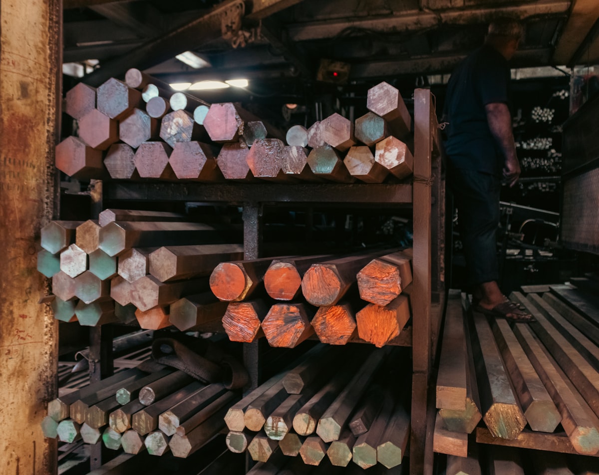 An industrial stockyard, featuring stacked hexagonal and circular metal bars on shelves. The varied ends of the bars display a spectrum of colors—from rust-red to oxidized green—creating a dense, repetitive pattern and geometric complexity. A worker on the right adds a sense of human scale to the heavy inventory. This texture is highly useful for engineering firm advertisements, as a conceptual background representing raw material wealth, or in editorial content about manufacturing and global supply chains. related to Discover the Elegance of Rod Iron Bed Frames