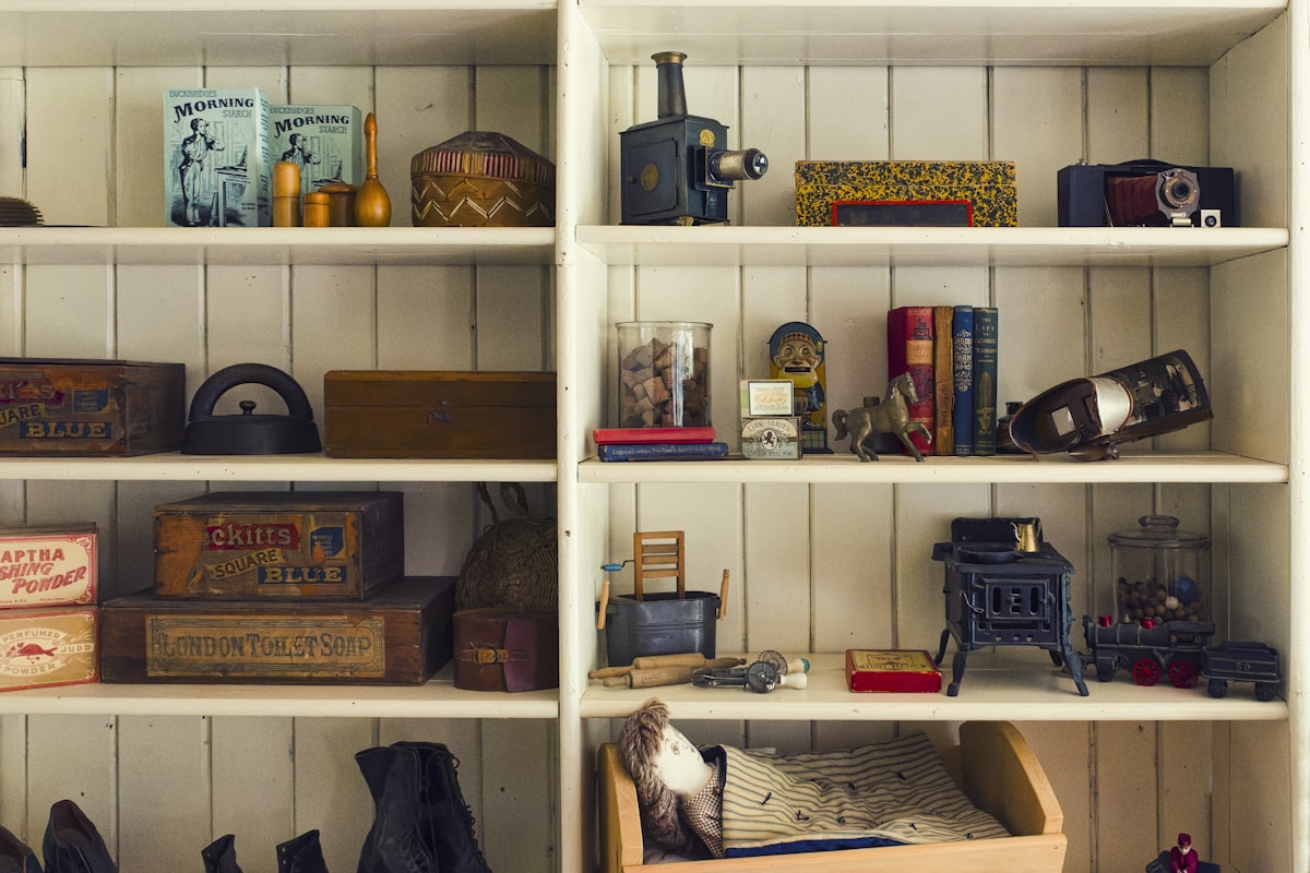 Antique object Collection Displayed On White Shelves - The image shows a collection of vintage toys and household items displayed on white wooden shelves. The collection includes an antique projector, a toy stove, a miniature bed with bedding, and various boxes of old-fashioned products. related to The Ultimate Guide to White Wooden Beds