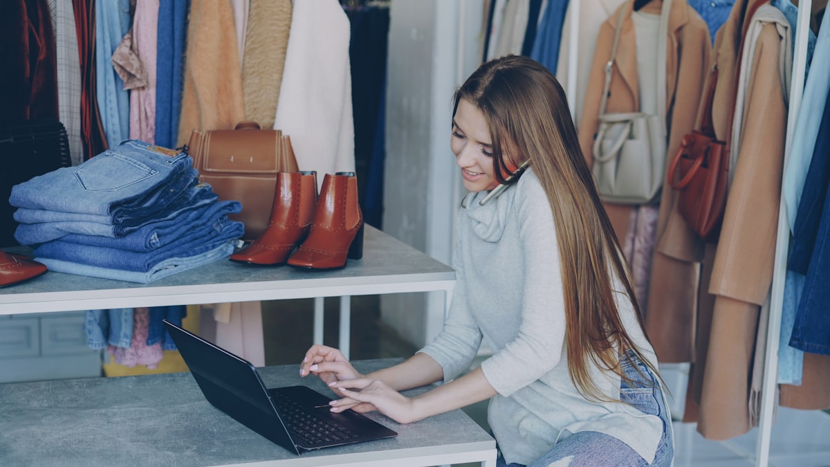 Attractive young businesswoman is sitting and talking on mobile phone while working with laptop in her women's clothing store. Colourful trendy garments, shoes and bags in background. related to Exploring Retail Options in Victor, NY