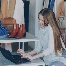 Attractive young businesswoman is sitting and talking on mobile phone while working with laptop in her women's clothing store. Colourful trendy garments, shoes and bags in background. related to Exploring Retail Options in Victor, NY