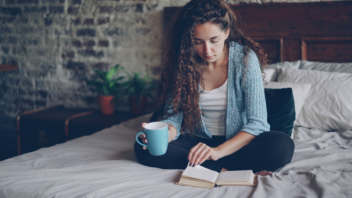 Attractive young woman with dark curly hair is reading book turning page and enjoying literature sitting on bed with cup of tea. Relaxed people, hobby and modern houses concept. related to A Cozy Escape: The Best Bed and Breakfasts in the Poconos