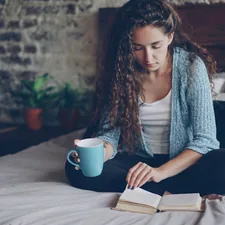 Attractive young woman with dark curly hair is reading book turning page and enjoying literature sitting on bed with cup of tea. Relaxed people, hobby and modern houses concept. related to A Cozy Escape: The Best Bed and Breakfasts in the Poconos