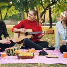 Charming young woman is playing the guitar sitting on blanket with friends on picnic, girls and guys are clapping hands and listening to music. Fun, millennials and nature concept. related to Transform Your Bedroom with Musical Themes