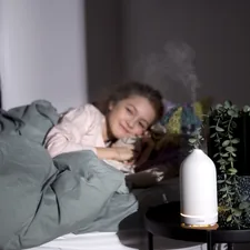 Girl smiling while resting on a bed with a white volant diffuser on her bedside table. related to The Ultimate Guide to Choosing a Bed Bath & Beyond Humidifier for Your Home