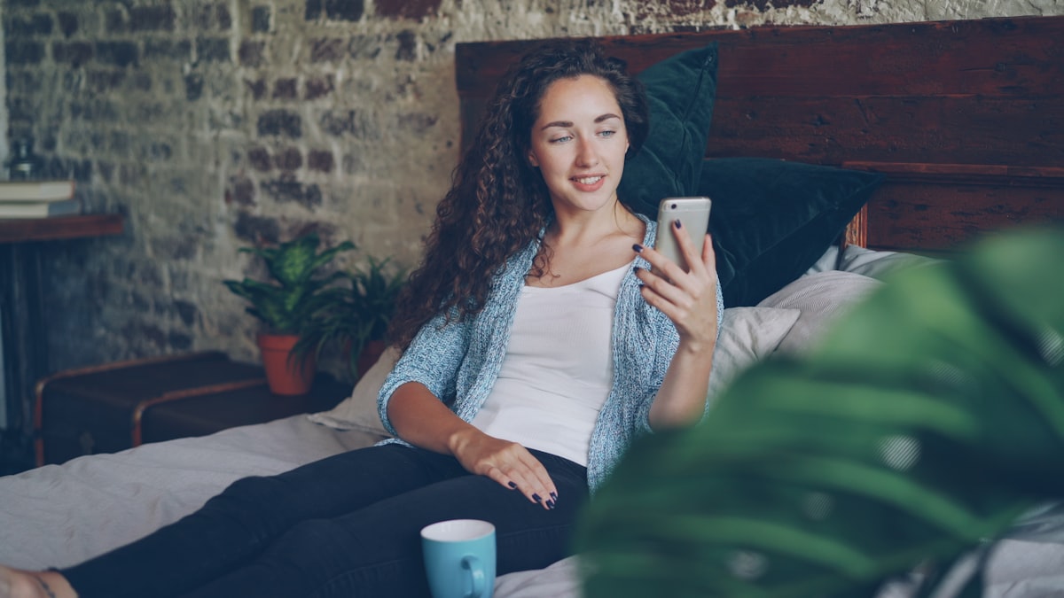 Good-looking woman is talking to friend online using modern smartphone making video call, she is happy and excited. Young lady is sitting on bed at home wearing casual clothes. related to Maximize Space with Innovative Wall Bed Solutions