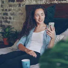 Good-looking woman is talking to friend online using modern smartphone making video call, she is happy and excited. Young lady is sitting on bed at home wearing casual clothes. related to Maximize Space with Innovative Wall Bed Solutions
