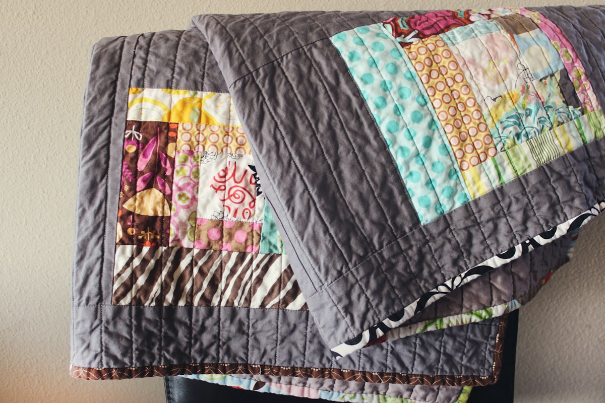Handmade colorful quilt hangs over a chair, in colors of gray, blue, pink, brown and yellow with abstract designed quilt blocks. related to The Cozy Conundrum: Coverlet vs Quilt