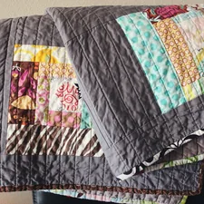 Handmade colorful quilt hangs over a chair, in colors of gray, blue, pink, brown and yellow with abstract designed quilt blocks. related to The Cozy Conundrum: Coverlet vs Quilt