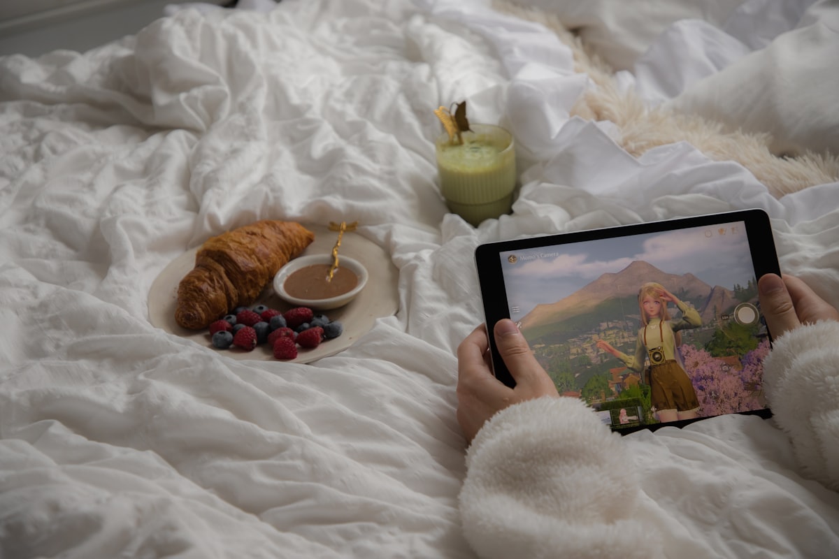 Hands holding a tablet playing 'Infinity Nikki', with a relaxing breakfast in bed situation featuring a matcha latte & croissant in the background. related to Beyond Pancakes: Elevating Your Breakfast in Bed Experience