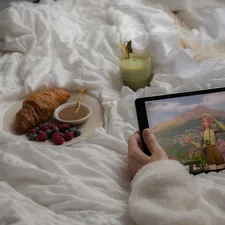 Hands holding a tablet playing 'Infinity Nikki', with a relaxing breakfast in bed situation featuring a matcha latte & croissant in the background. related to Beyond Pancakes: Elevating Your Breakfast in Bed Experience