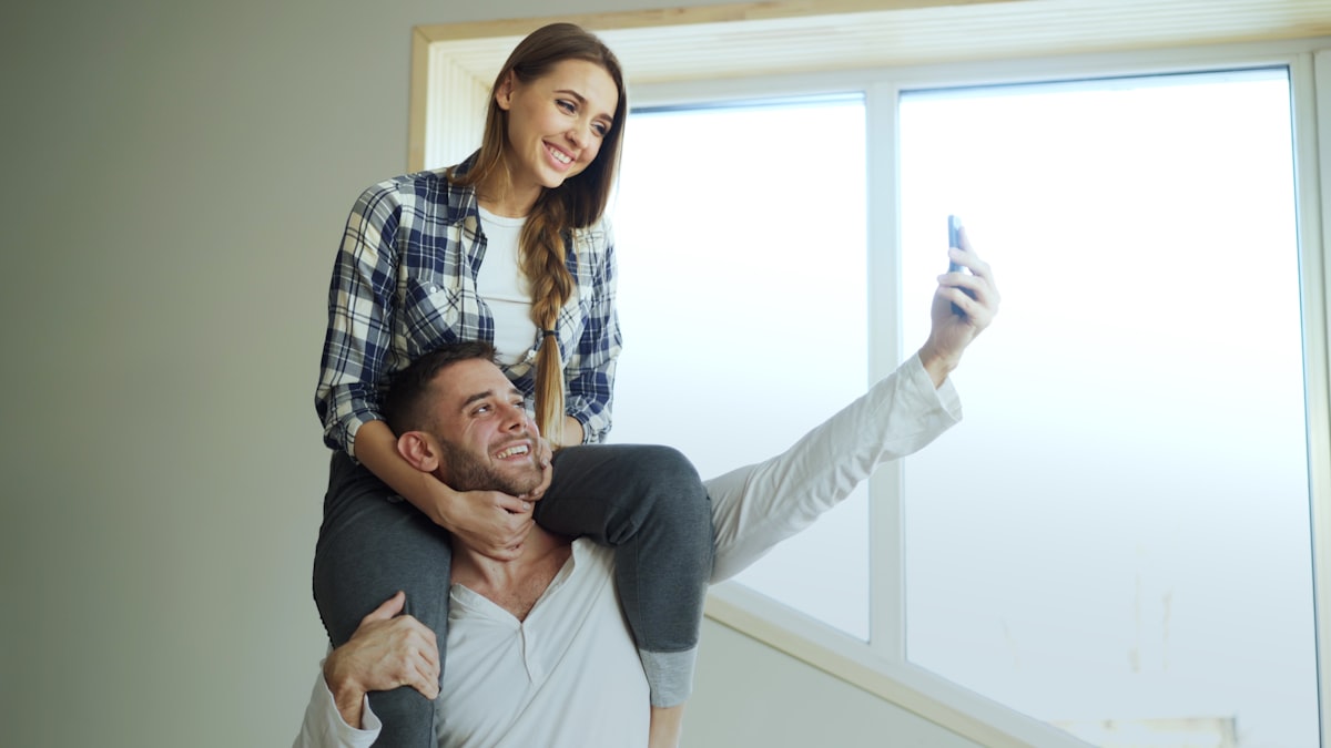 Happy young couple having fun and talking online video call in the kitchen. Girl is sitting on boyfriend's neck while he holding smartphone related to Elevate Your Home's First Impression with Walmart Entryway Furniture