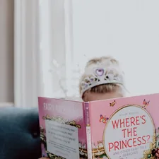 Little girl wearing a princess crown reading a "Where's the Princess" book related to Finding the Perfect Princess Crib Bedding Set for Your Little Royal