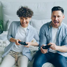 Man and woman playing video game sitting on bed in apartment having fun related to Transform Your Gaming Space: The Ultimate Guide to Gaming Bed Sheets