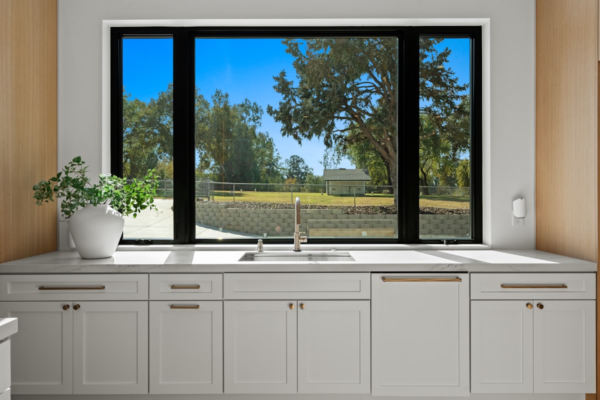 Modern kitchen sink with white shaker cabinetry, brass hardware, and large black-framed picture window overlooking greenery. Bright contemporary home interior featuring marble countertops, natural light, and clean minimalist design. High-end residential kitchen with timeless finishes and warm wood accents. related to Kitchen Window Valance Ideas to Elevate Your Cooking Space