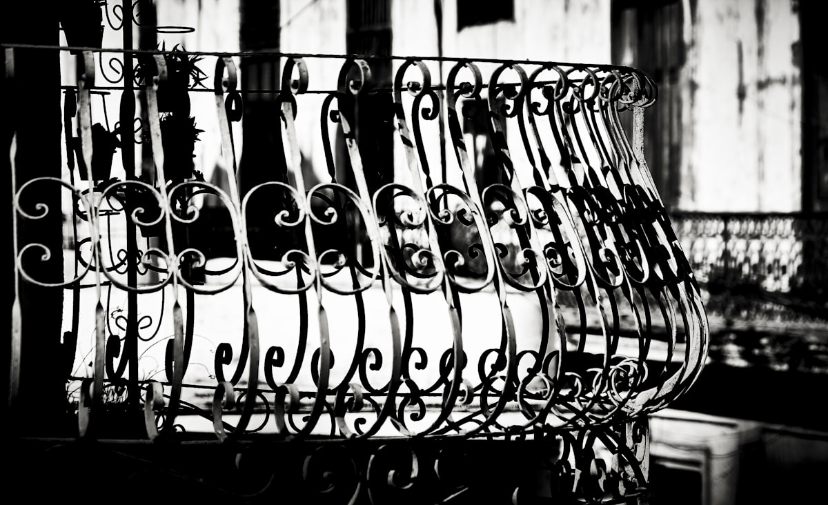 Ornate wrought iron balcony railing with scrollwork details, captured in high contrast black and white. The intricate curves and spirals reflect the craftsmanship of colonial-era architecture. related to Wrought Iron Bed Frames - Elevate Your Home's Style