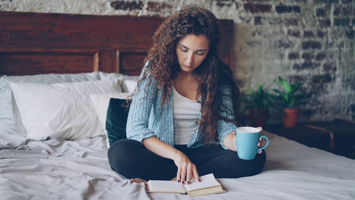 Pretty girl is reading book sitting on bed at home and holding cup with drink enjoying free time and resting. Young people, hobby and modern interior concept. related to A Cozy Escape: The Best Bed and Breakfasts in the Poconos