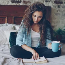 Pretty girl is reading book sitting on bed at home and holding cup with drink enjoying free time and resting. Young people, hobby and modern interior concept. related to A Cozy Escape: The Best Bed and Breakfasts in the Poconos