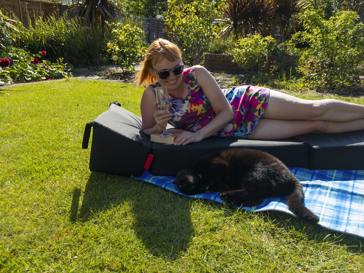 Smiling woman wearing sunglasses in a short, colourful summer dress lying in the sun on a padded lounger and a blue picnic blanket in the garden holding a glass of champagne with her cat lying next to her related to Choosing the Perfect Dog Ramp for Your Bed: A Comprehensive Guide
