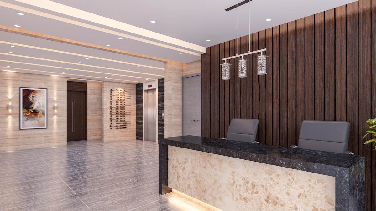 The image is a wide shot of a spacious and modern building lobby. A large, dark-topped reception desk with a marbled base dominates the right foreground. Two gray office chairs are tucked neatly behind it, and the wall behind the desk is covered in vertical wooden slats. A contemporary chandelier hangs above the desk. To the left, a set of elevators with brushed metal doors and a sleek directory panel are visible. The walls are a mix of light-toned polished stone and what looks like a white wall with an abstract painting. The floor is a reflective, light-colored tile with a polished finish. Th related to Welcome Home: Elevate Your Space with These Entryway Decorating Ideas