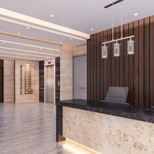 The image is a wide shot of a spacious and modern building lobby. A large, dark-topped reception desk with a marbled base dominates the right foreground. Two gray office chairs are tucked neatly behind it, and the wall behind the desk is covered in vertical wooden slats. A contemporary chandelier hangs above the desk. To the left, a set of elevators with brushed metal doors and a sleek directory panel are visible. The walls are a mix of light-toned polished stone and what looks like a white wall with an abstract painting. The floor is a reflective, light-colored tile with a polished finish. Th related to Welcome Home: Elevate Your Space with These Entryway Decorating Ideas