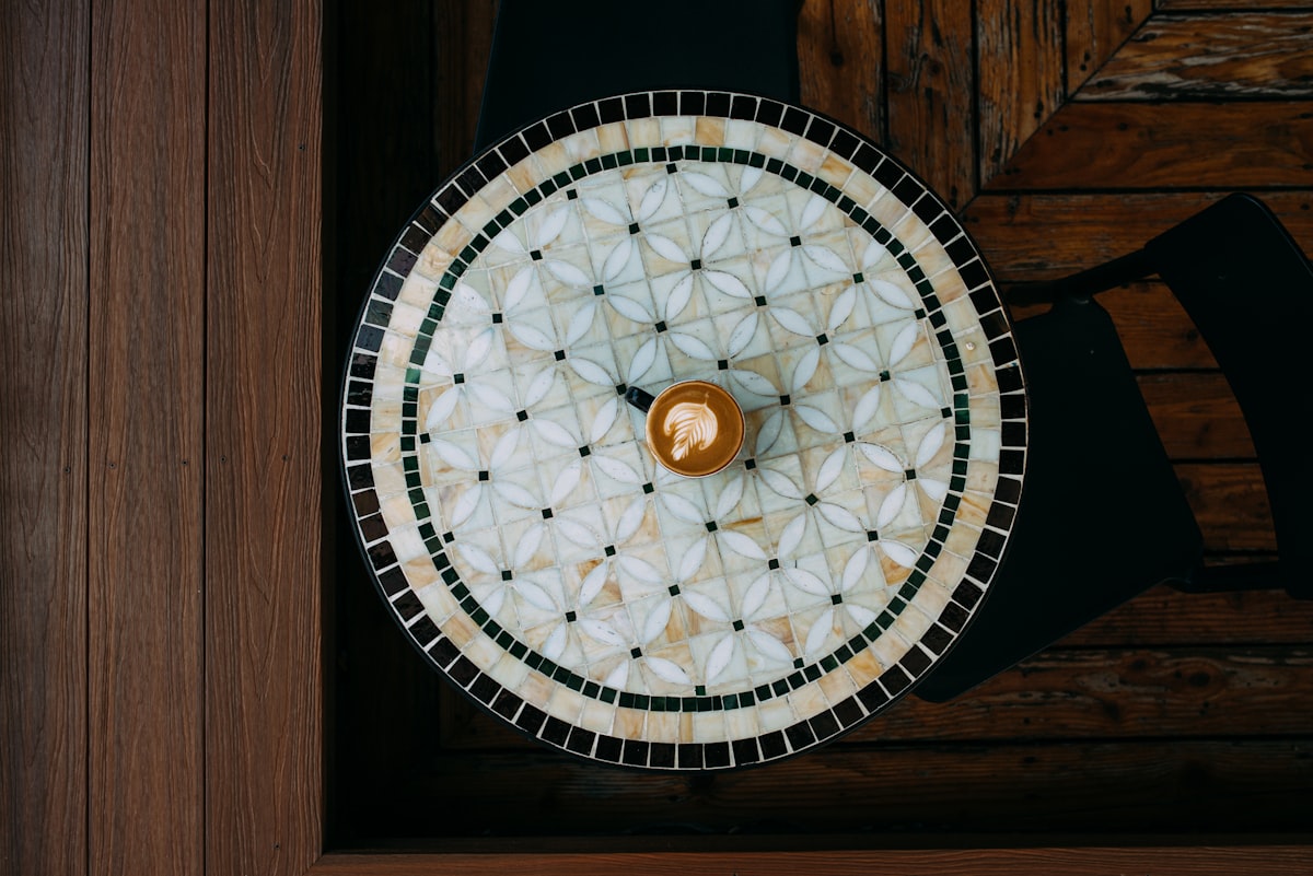 Top-down view of a small latte cup resting on a round mosaic tile table with warm wooden surroundings in a cafe. related to Elevate Your Entryway With a Stunning Round Entryway Table