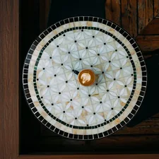 Top-down view of a small latte cup resting on a round mosaic tile table with warm wooden surroundings in a cafe. related to Elevate Your Entryway With a Stunning Round Entryway Table