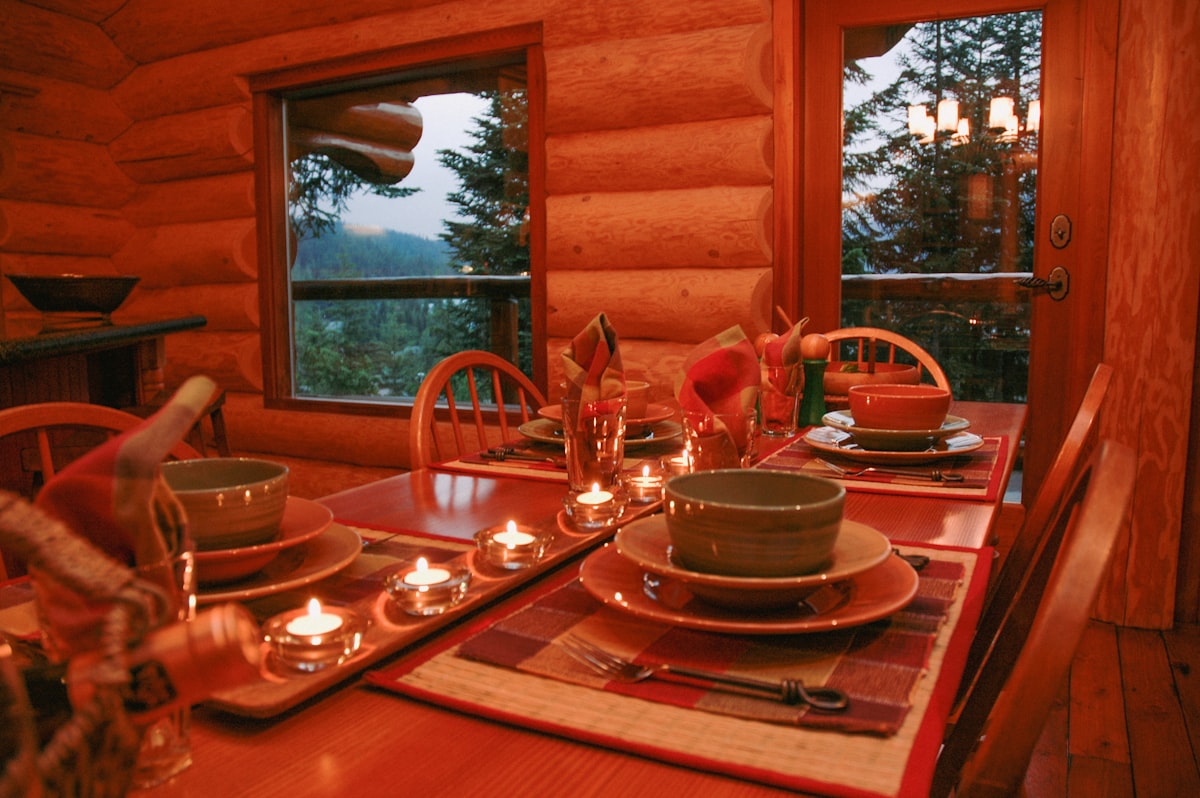 Wooden dining table with bowls and plates. Ready for dinner with candles lit, view of trees out the window. related to The Ultimate Guide to Log Cabin Bedding: Creating the Perfect Rustic Retreat