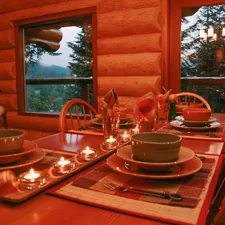 Wooden dining table with bowls and plates. Ready for dinner with candles lit, view of trees out the window. related to The Ultimate Guide to Log Cabin Bedding: Creating the Perfect Rustic Retreat