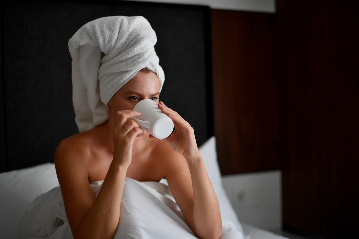 Young blonde woman with a white towel wrapped around her head drinking hot coffee in bed during her morning routine. related to Sleep Better with Bed Head Products: Expert Reviews and Buying Guide